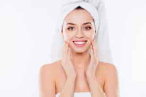 Portrait of stunning woman in towel and turban on head enjoying touching her perfect face skin with fingers isolated on white background