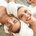 Two young beautiful smiling women in white bathrobes and towels on head.